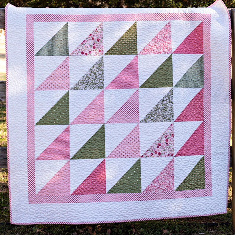 May include: A handmade quilt with a geometric design. The quilt features alternating triangles in shades of pink and green, set against a white background. A pink dotted border frames the quilt, which is likely made of cotton.