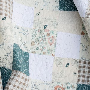 May include: A quilted blanket featuring a patchwork design of square fabric panels. The pattern includes solid white squares with a dotted texture, dark green squares with white floral prints, and cream squares with floral and moth motifs.
