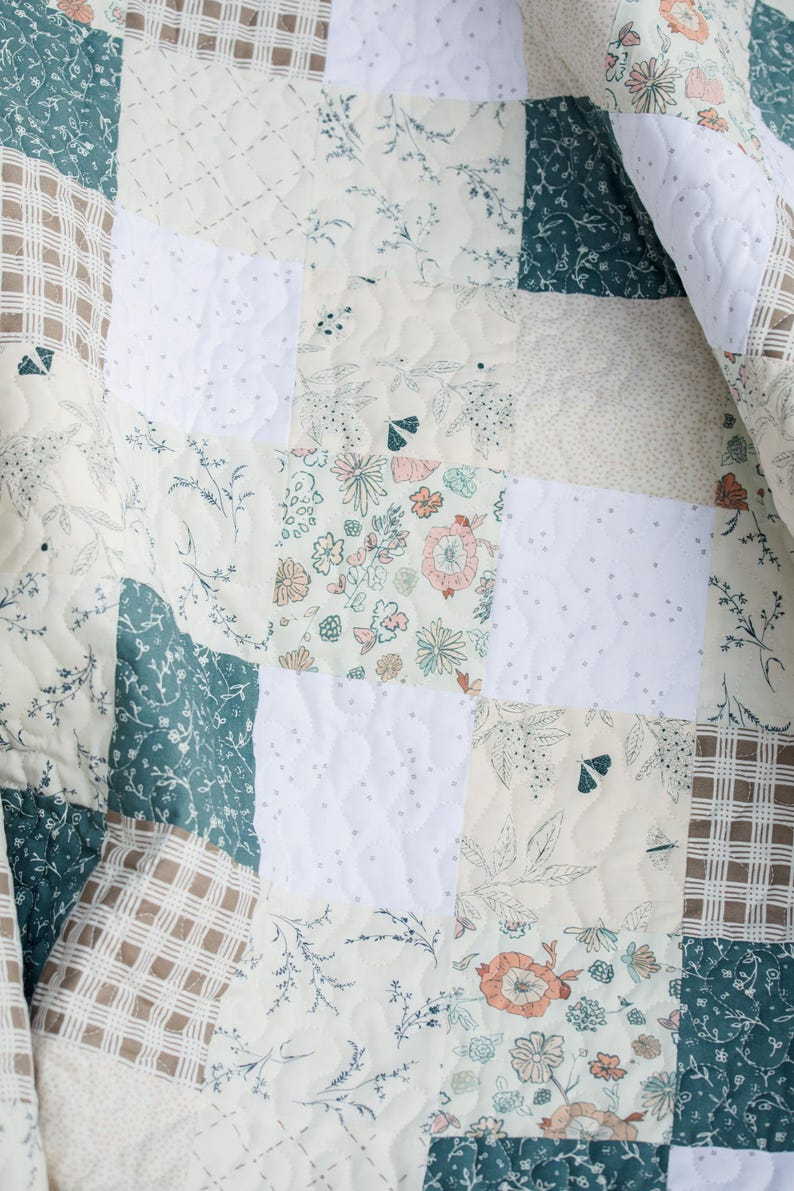 May include: A quilted blanket featuring a patchwork design of square fabric panels. The pattern includes solid white squares with a dotted texture, dark green squares with white floral prints, and cream squares with floral and moth motifs.