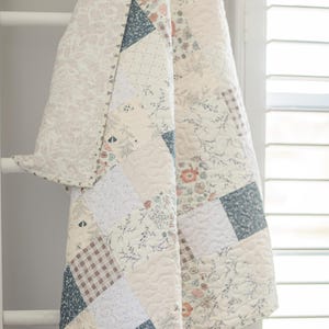 May include: A patchwork quilt featuring a variety of square fabric panels in muted tones of blue, cream, and soft pink. The design includes floral patterns, small-scale botanical prints, and a brown and white gingham check, all finished with stitching.