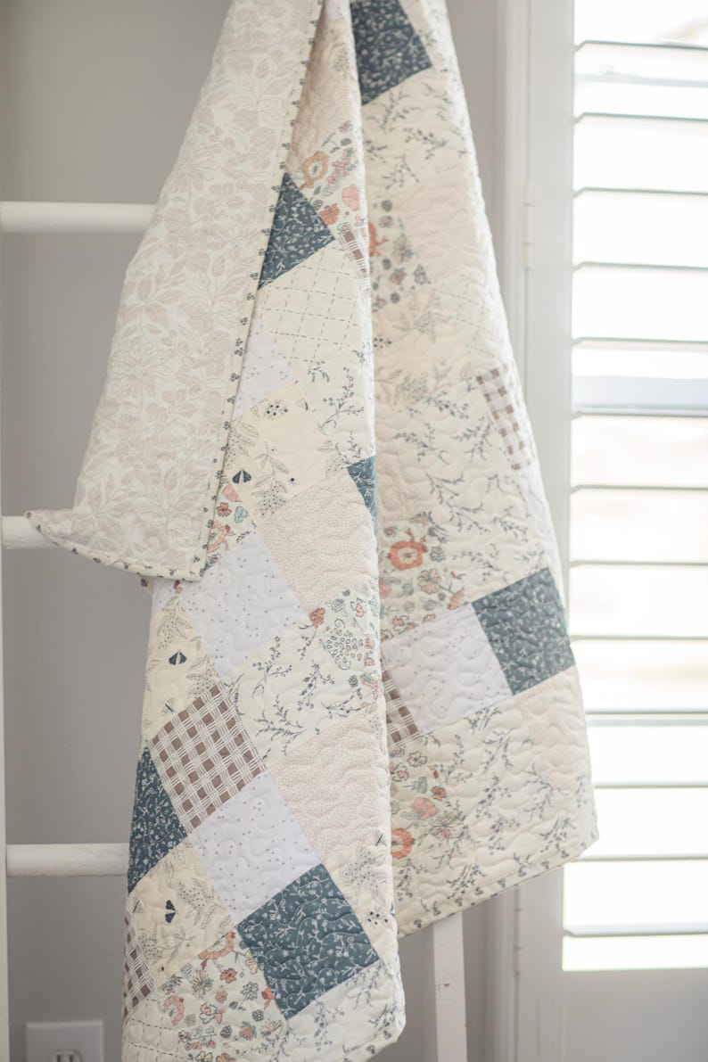 May include: A patchwork quilt featuring a variety of square fabric panels in muted tones of blue, cream, and soft pink. The design includes floral patterns, small-scale botanical prints, and a brown and white gingham check, all finished with stitching.