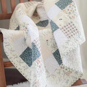 May include: A patchwork quilt featuring a variety of square fabric blocks in shades of cream, sage green, and soft brown. The quilt displays patterns including floral prints, small gingham checks, and solid textures with an overall quilted stitch.