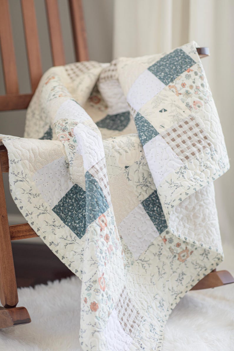 May include: A patchwork quilt featuring a variety of square fabric blocks in shades of cream, sage green, and soft brown. The quilt displays patterns including floral prints, small gingham checks, and solid textures with an overall quilted stitch.
