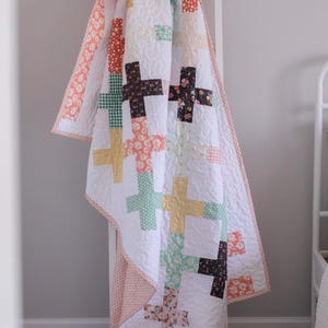 Puede incluir: Un edredón blanco con un patrón de cruces en varios colores, incluyendo amarillo, marrón y melocotón. El edredón está colgado sobre una escalera blanca, mostrando su diseño y textura. El borde del edredón presenta un patrón floral melocotón.