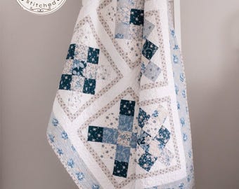 May include: A quilt featuring a patchwork design in shades of navy blue, light blue, and white with floral and geometric patterns. The fabric is stitched in a grid and diamond layout. A circular logo with the text gathered stitched is visible.