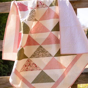 May include: A handmade quilt with a patchwork design featuring triangles in shades of pink, green, and white. The quilt has a red and white gingham border and is draped over a wooden fence. The quilt is approximately 152 cm by 178 cm.