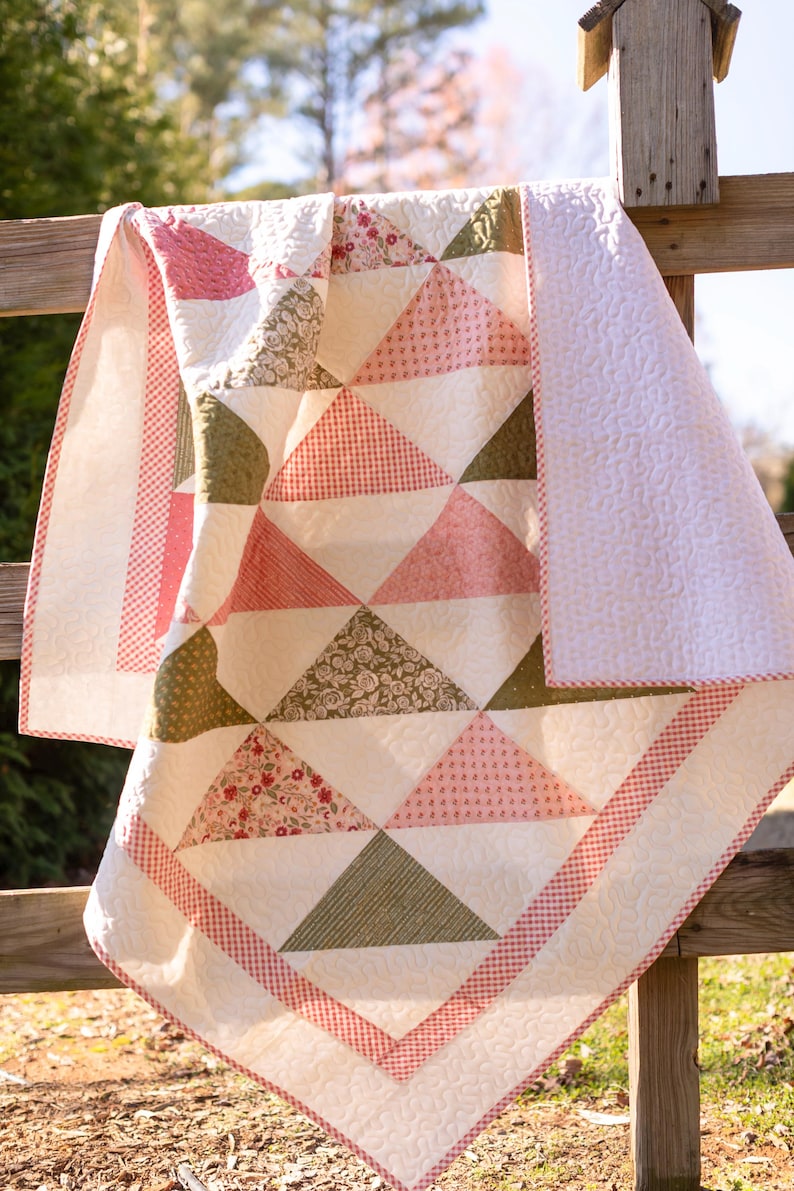 May include: A handmade quilt with a patchwork design featuring triangles in shades of pink, green, and white. The quilt has a red and white gingham border and is draped over a wooden fence. The quilt is approximately 152 cm by 178 cm.