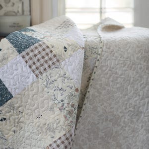 May include: A patchwork quilt featuring a variety of square fabric blocks in shades of cream, white, muted blue, and brown. The design includes floral patterns, small-scale botanical prints, and brown-and-white gingham checks with stippled quilting.