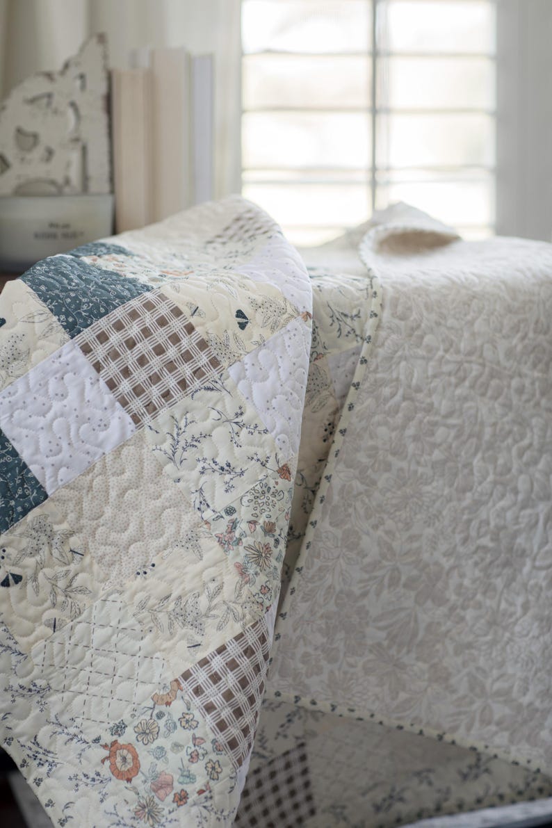 May include: A patchwork quilt featuring a variety of square fabric blocks in shades of cream, white, muted blue, and brown. The design includes floral patterns, small-scale botanical prints, and brown-and-white gingham checks with stippled quilting.
