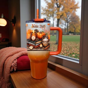 May include: An orange tumbler with a handle and straw featuring a graphic print of a pumpkin, a lit candle, a stack of books, and a mug with the text Hello Fall. The tumbler has a gradient design transitioning from a white top to an orange base.