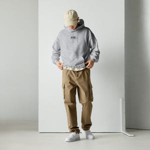 May include: A person wearing a grey hoodie with the word "graph" embroidered on the chest, paired with khaki cargo trousers, a beige baseball cap, and white trainers. The outfit is casual and comfortable.