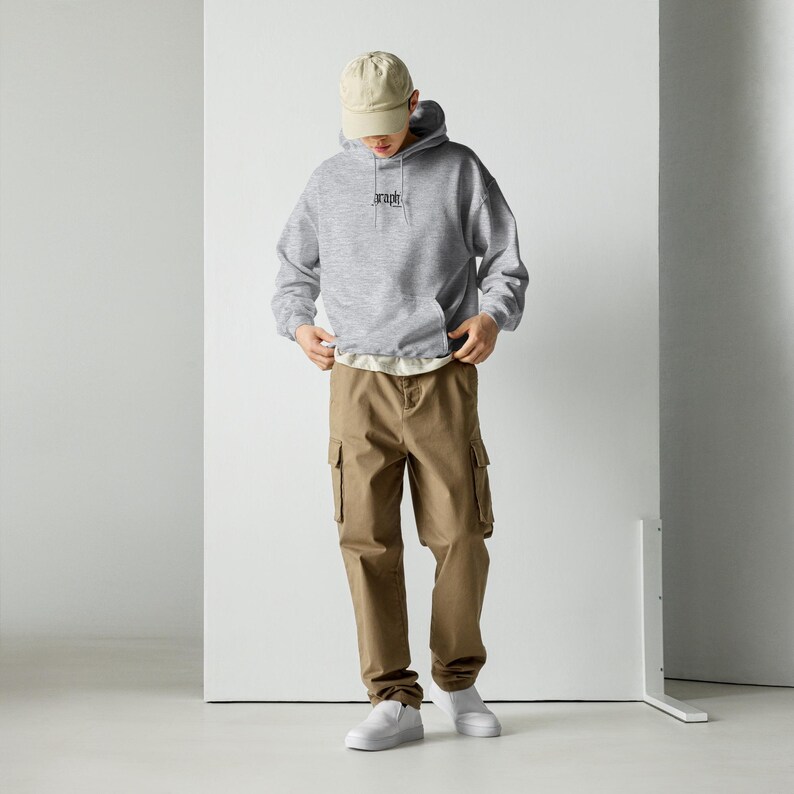 May include: A person wearing a grey hoodie with the word "graph" embroidered on the chest, paired with khaki cargo trousers, a beige baseball cap, and white trainers. The outfit is casual and comfortable.