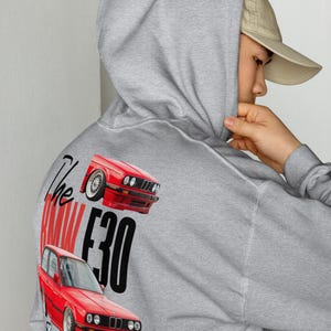 May include: A grey hoodie with a red car graphic and the text "The BMW E30". The design features two red car illustrations. The hoodie has a hood and a front pocket.