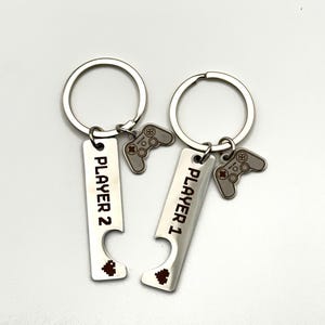 May include: Two silver keychains with the words "PLAYER 1" and "PLAYER 2" engraved on them. Each keychain has a small, silver controller charm and a bottle opener design at the bottom. The keychains are connected to a silver ring.