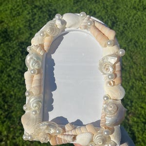 Könnte beinhalten: Weißer Bilderrahmen, verziert mit verschiedenen Muscheln und Kunstperlen. Der Rahmen hat eine gewölbte Oberseite und ist von einer Vielzahl von Muscheln in Weiß-, Creme- und Beigetönen umgeben. Kleine, runde Perlen sind zwischen den Muscheln verstreut.