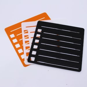 May include: Three stencils in orange, white, and black, each resembling a notepad page. The stencils have horizontal lines and a vertical column of small squares on the left side, creating a lined paper effect. The stencils are overlapping.