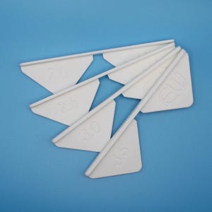 May include: A set of four white plastic corner gauges arranged in a fan shape. Each gauge features a triangular head with a raised edge and is embossed with numerical size markings 2.0, 2.5, 3.0, and 3.5, along with the letters EW.