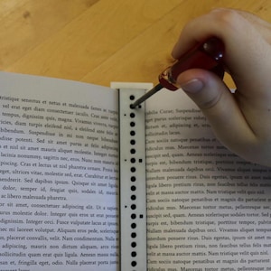 May include: A red screwdriver is being used to adjust a screw in a white, perforated strip. The strip is next to a book with Latin text. The image focuses on the tool and the action of adjusting the screw.