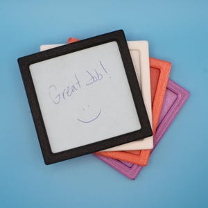 May include: A stack of four square dry-erase boards with textured frames in black, white, orange, and purple. The top black-framed board features the handwritten text Great Job! and a smiley face drawn in blue ink on the white writing surface.