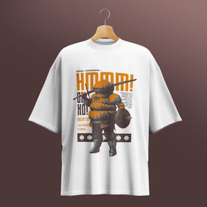 May include: White t-shirt featuring a cartoon knight in orange and grey armour, holding a sword and shield. The shirt has the text "Hmm... Oh Ho!" in orange, with more text below. The t-shirt is suspended from a wooden hanger.