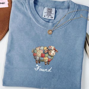 May include: A blue t-shirt featuring a central embroidered graphic of a patchwork sheep with colorful floral and geometric patterns. Below the sheep, the word Found is embroidered in white script. The shirt has a crew neckline and a tag.