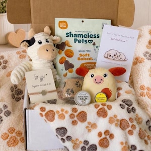May include: A gift box filled with dog-themed items. Includes a plush cow, a pig toy, a blanket with paw prints, treats, a card that says "Sorry You're Feeling Ruff", and a paw and nose balm. The box is open, revealing the contents.