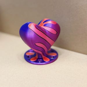 May include: A heart-shaped decorative object with a base. The heart is purple with pink and orange stripes. The base is a dark purple with orange accents. The object is 3D printed.