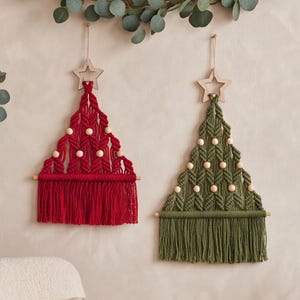 以下が含まれることがあります： クリスマスツリーの形をしたマクラメ編みの壁掛け2点。1点はダークレッド、もう1点はオリーブグリーンです。それぞれ上部に木製の星、編み込み模様の中に明るい色の木製ビーズがあしらわれ、下部には木製の棒から吊り下げられたフリンジが付いています。