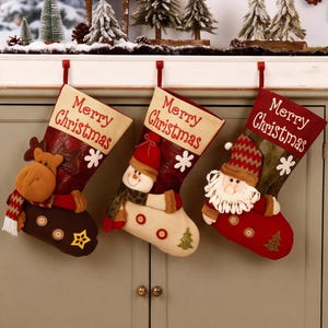 Puede incluir: Tres calcetines de Navidad colgados en fila, cada uno con el texto Merry Christmas en la parte superior. El calcetín izquierdo muestra un reno, el central un muñeco de nieve y el derecho un Santa Claus, todos con botones decorativos.