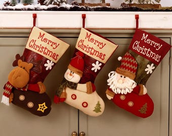 以下が含まれることがあります： Merry Christmasの文字が上部に刺繍された3つのクリスマスストッキングが並んで吊るされています。左のストッキングにはトナカイ、中央には雪だるま、右にはサンタクロースのぬいぐるみが付いており、それぞれ装飾用のボタンがあしらわれています。