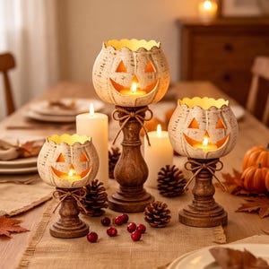 May include: Three off-white, textured pumpkin-shaped candle holders with carved jack-o'-lantern faces, each resting on a dark brown wooden pedestal base tied with a thin twine bow. Each holder contains a glowing tea light candle.
