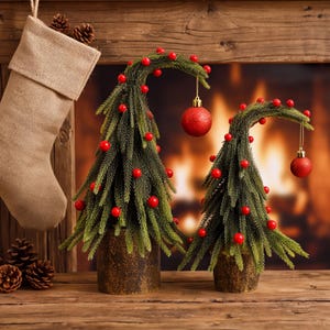 Puede incluir: Dos árboles de pino artificiales verdes con puntas curvas, decorados con pequeñas bayas rojas y un adorno rojo brillante colgante en cada uno. Los árboles están en macetas cilíndricas marrones texturizadas, junto a un calcetín de arpillera.