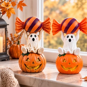 May include: Two Halloween figurines featuring white ghosts sitting atop orange jack-o'-lanterns. Each ghost holds a large, striped orange and purple candy above its head. The ghosts have black eyes, mouths, and pink cheeks, while the pumpkins have carved faces.