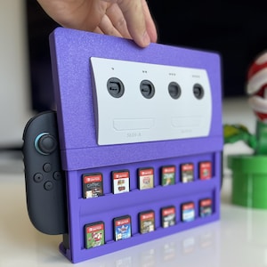 Op de afbeelding: Een paars gameconsole-dock met een wit voorpaneel met vier controllerpoorten en twee geheugenkaartsleuven met het label SLOT-A en SLOT-B. Het dock bevat een zwarte controller aan de zijkant en twee rijen met diverse gamecartridges.