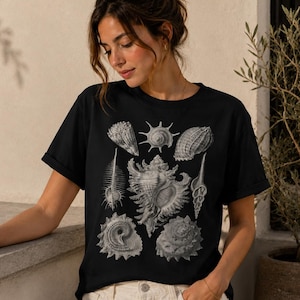 Puede incluir: Una camiseta negra de manga corta con un estampado gráfico monocromático detallado de varias conchas marinas dispuestas en grupo. El diseño incluye múltiples especies de conchas en tonos blancos, grises y negros en la parte frontal.
