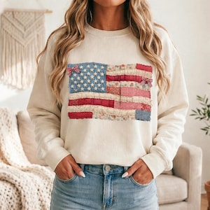 Könnte beinhalten: Cremefarbenes Sweatshirt mit einem Patchwork-Design der amerikanischen Flagge. Die Flagge enthält blauen Denim mit weißen Sternen, rote Streifen und florale und Vichy-Stoffakzente. Kleine rot-weiße Schleifen sind an der Flagge befestigt.