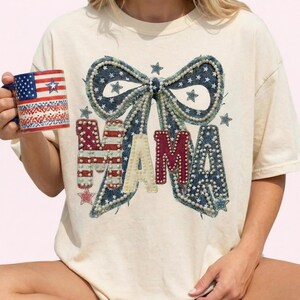 May include: Cream-colored t-shirt with a denim bow and the word "MAMA" in red, cream, and denim. The bow is adorned with pearls and stars. A mug with an American flag design is held in the hand.