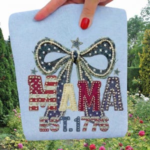 May include: Light blue decorative square with a patriotic bow and the word "MAMA" in red, white, and blue. The text "EST. 1776" is also displayed. The design is embellished with pearl-like beads and star accents.
