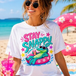 以下が含まれることがあります： サングラスをかけてピンクの花柄の浮き輪でリラックスしている緑色のワニのグラフィックプリントが施された、白い半袖Tシャツ。ワニの上には「STAY SNAPPY」という文字が鮮やかなピンクとティールブルーでプリントされています。
