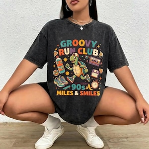 May include: Dark gray t-shirt with a colorful graphic. The graphic features a running turtle, a cassette tape, sneakers, and the text "GROOVY RUN CLUB 90s MILES & SMILES". The shirt has short sleeves and a crew neck.