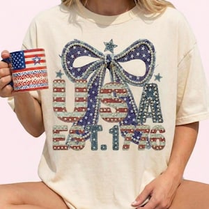 May include: Cream-colored t-shirt with a large USA graphic in red, white, and blue, featuring a bow and stars. The text "EST. 1776" is below the letters. A mug with an American flag design is held in the hand.