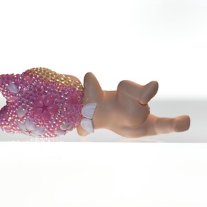 K&ouml;nnte beinhalten: Eine Figur eines Babys, das liegt, mit einem rosa-goldenen, mit Juwelen besetzten Kopfschmuck. Die Haut des Babys ist hellbraun und es tr&auml;gt wei&szlig;e Windel&uuml;berhosen. Die Figur befindet sich auf einer wei&szlig;en Oberfl&auml;che.