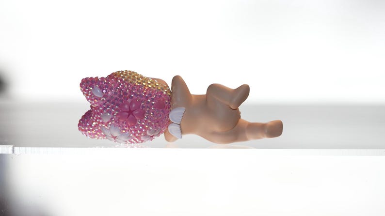 K&ouml;nnte beinhalten: Eine Figur eines Babys, das liegt, mit einem rosa-goldenen, mit Juwelen besetzten Kopfschmuck. Die Haut des Babys ist hellbraun und es tr&auml;gt wei&szlig;e Windel&uuml;berhosen. Die Figur befindet sich auf einer wei&szlig;en Oberfl&auml;che.