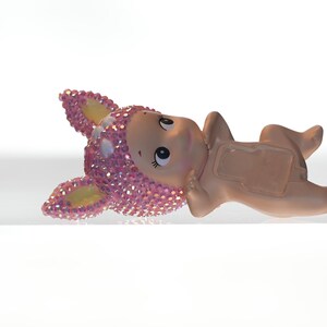 K&ouml;nnte beinhalten: Keramikfigur eines liegenden Babys mit einem rosa, mit Juwelen besetzten Tierkopf. Die Figur hat einen hellen Hautton und ist vor einem wei&szlig;en Hintergrund platziert. Der Tierkopf hat gelbe Akzente.