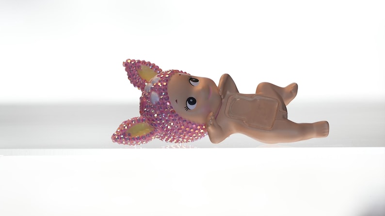 K&ouml;nnte beinhalten: Keramikfigur eines liegenden Babys mit einem rosa, mit Juwelen besetzten Tierkopf. Die Figur hat einen hellen Hautton und ist vor einem wei&szlig;en Hintergrund platziert. Der Tierkopf hat gelbe Akzente.