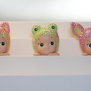 K&ouml;nnte beinhalten: Drei kleine Figuren mit l&auml;chelnden Gesichtern, die jeweils einen Hut mit Tiermotiv tragen. Die H&uuml;te sind mit glitzernden Kristallen in Rosa, Gelb und Gr&uuml;n verziert. Die Figuren werden auf einer wei&szlig;en, abgestuften Oberfl&auml;che ausgestellt.