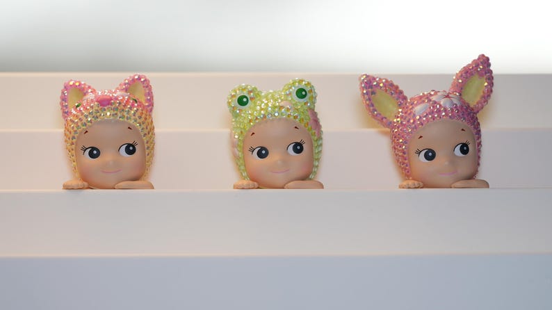 K&ouml;nnte beinhalten: Drei kleine Figuren mit l&auml;chelnden Gesichtern, die jeweils einen Hut mit Tiermotiv tragen. Die H&uuml;te sind mit glitzernden Kristallen in Rosa, Gelb und Gr&uuml;n verziert. Die Figuren werden auf einer wei&szlig;en, abgestuften Oberfl&auml;che ausgestellt.