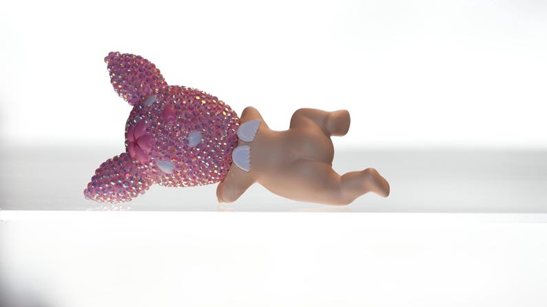 K&ouml;nnte beinhalten: Eine rosa Hasenfigur mit kristallartiger Textur auf Kopf und Ohren, gepaart mit einer hellh&auml;utigen Babyfigur. Der Hase liegt auf der Seite, die Babyfigur ist davor positioniert.