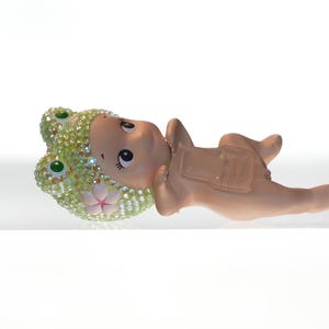 K&ouml;nnte beinhalten: Eine skurrile Figur eines Babys in einem Froschkost&uuml;m, das auf dem R&uuml;cken liegt. Die Froschhaube ist mit glitzernden Strasssteinen verziert. Die Haut des Babys ist hellbraun und das Kost&uuml;m hat eine passende braune Farbe.