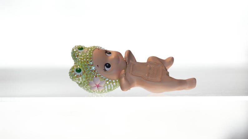 K&ouml;nnte beinhalten: Eine skurrile Figur eines Babys in einem Froschkost&uuml;m, das auf dem R&uuml;cken liegt. Die Froschhaube ist mit glitzernden Strasssteinen verziert. Die Haut des Babys ist hellbraun und das Kost&uuml;m hat eine passende braune Farbe.